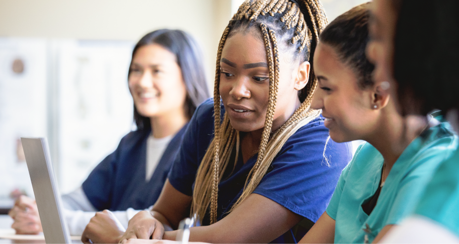 Um grupo diversificado de mulheres vestidas com batas médicas sentadas juntas à mesa, destacando a união e a cooperação num ambiente médico.