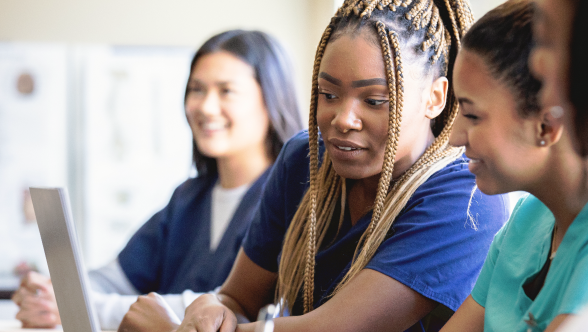 Um grupo diversificado de mulheres vestidas com batas médicas sentadas juntas à mesa, destacando a união e a cooperação num ambiente médico.
